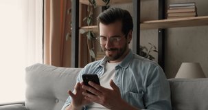 Happy young 30s handsome man in eyeglasses resting on cozy sofa with smartphone in hands, enjoying shopping online, playing mobile game, communicating distantly in social network, reading media news.