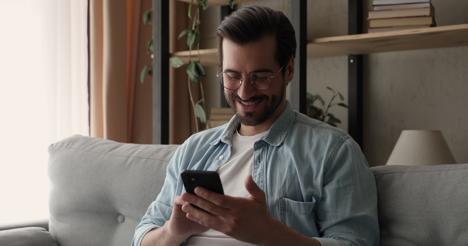 Happy young 30s handsome man in eyeglasses resting on cozy sofa with smartphone in hands, enjoying shopping online, playing mobile game, communicating distantly in social network, reading media news.