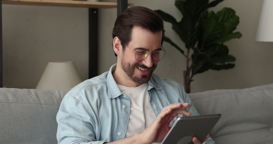 Smiling relaxed carefree young man in glasses using digital tablet apps, sitting on couch, enjoying shopping in internet store choosing goods, communicating distantly with friends or web surfing. Royalty-Free Stock Footage #1075713113