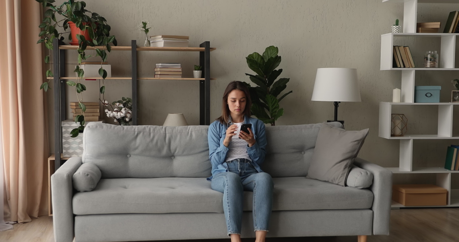 Happy emotional young woman sitting on sofa, reading message or email on smartphone, feeling excited of getting good news, win notification, commercial offer, celebrating online success at home. Royalty-Free Stock Footage #1075713128
