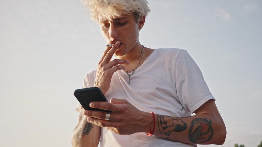 Portrait of young fashionable blonde with tattoos smokes a cigarette and holds a smartphone in his hands against the background of the summer sky. Youth culture and bright appearance.