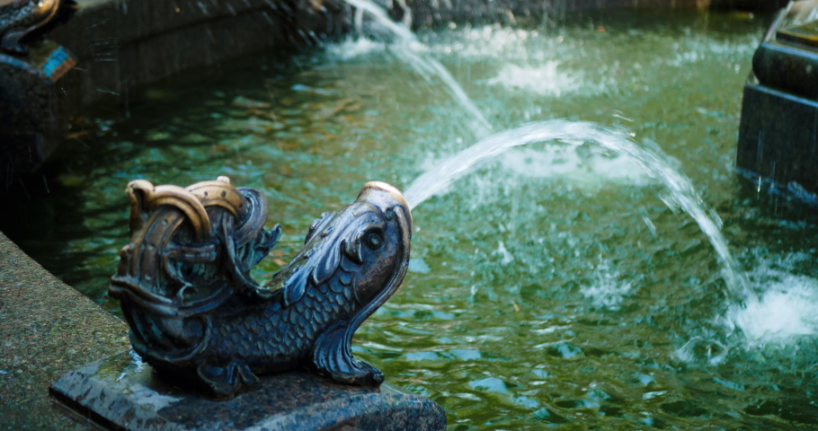Beautiful fountain in a city park in a modern style. A woman in a blue dress with flowers and a brown bag washes her hands under a stream of water from a fabulous copper fish. Water splash. Close Up