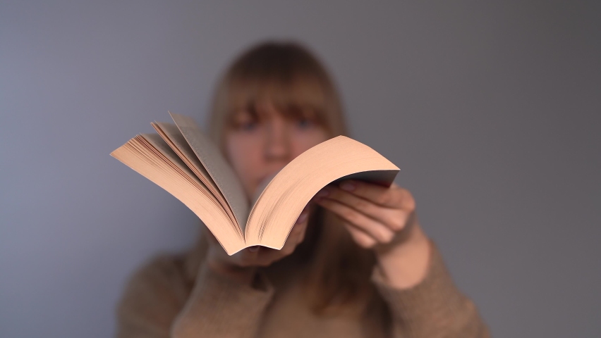 Young blonde university or college student girl dressed in beige sweater leafing through a book in front of camera on a gray background. Knowledge or educational concept. High quality 4k video