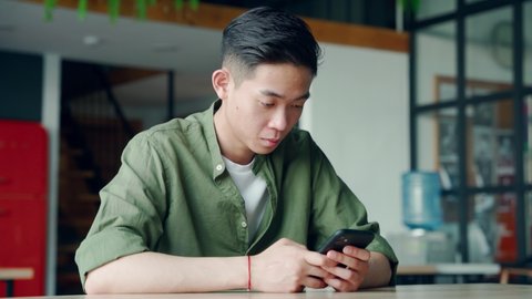 Young smart asian chinese happy student man using apps holding gadget cell phone at desk in office smiling texting. Online learning work education mobile technology concept.