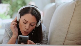 Video of motivated young woman listening and singingto music with smartphone while sitting on sofa at home.