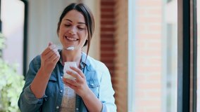 Video of beautiful businesswoman eating yogurt while standing near the windows looking at camera in living room at home.