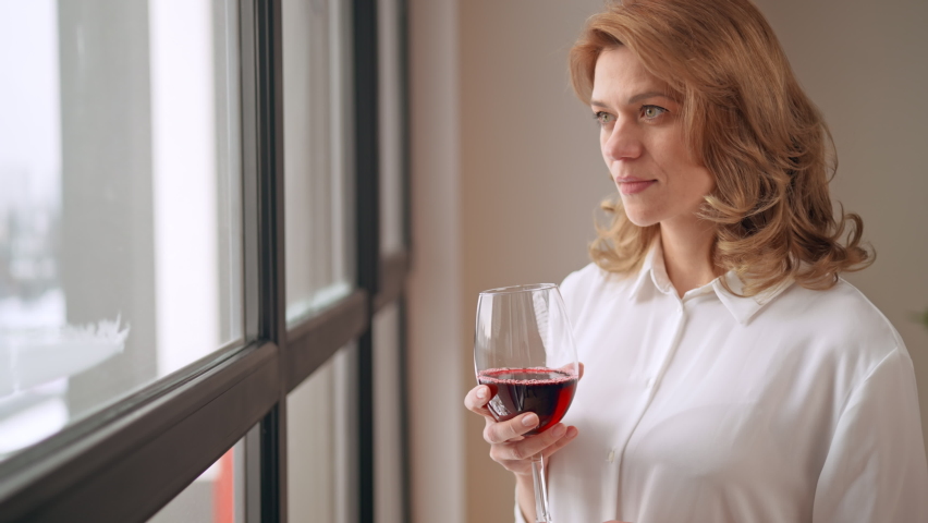 side view elegant middle aged woman in white shirt standing near window drinking red wine time for break and relax