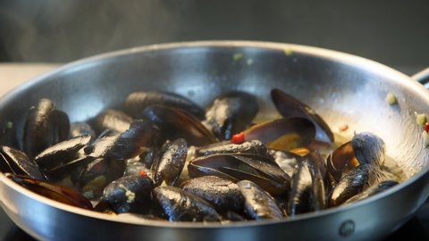 Cooking delicious fresh mussels on hot pan in luxury restaurant. Chef shakes and mixes mussels and greens on a grill. Traditional mediterranean cuisine. Slow motion scene.