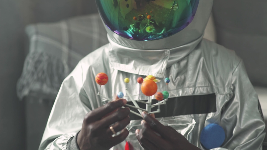 African man in an astronaut costume sitting on the floor in living room at home and playing with a toy model of the solar system, study of astronomy, man on self-isolation.