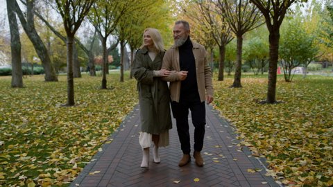 Charming elderly man and beautiful woman looking each other in autumn park. Happy married couple walking along foliage fall path. Smiling lovers enjoying together outdoors.
