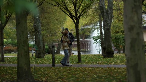 Happy couple in love having romantic walk with joy and fun at park. Wide shot of young lovers kissing in hugging in fall nature. Sweetheart girl and guy falling in love with fountain on background. 