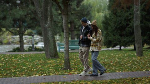 Smiling young lovers walking in beautiful autumn park. Wide shot of happy stylish girl and guy having walking with enjoyment in fall weather. Romantic couple dating outdoors.