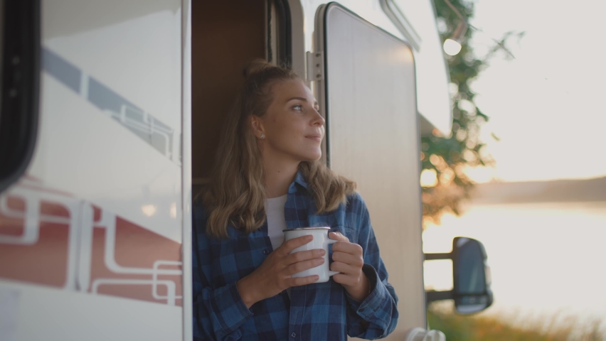 Caucasian female is resting in nature near the lake, standing near a mobile home and enjoying nature holding a mug of hot tea in her hands, summer vacation, woman near camping car, 4k slow motion.
