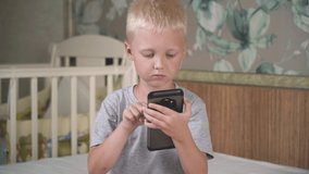 Cute cheerful little caucasian boy spending time on a social network or playing video games on a mobile phone sitting on a big bed. Lifestyle concept. Communication and games on a smartphone.