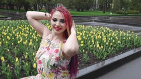 Awesome girl with rainbow braids and expressive glitter makeup. She sits and enjoys the warm spring weather in a green park with large tulip beds. UHD 4K 4x slow-motion video