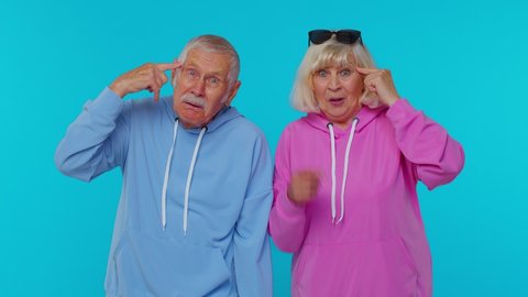 You are crazy, out of mind? Senior old retired grandfather grandmother pointing at camera, showing stupid gesture, blaming some idiot for insane plan on blue background. Mature man woman grandparents