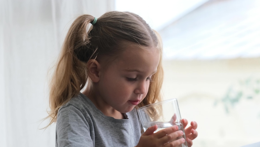 Little blonde girl in white dress drinks water from a glass indoors. Cute child is drinking a cup of water | Shutterstock HD Video #1075742561