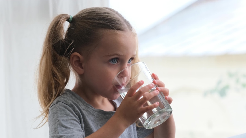 Little blonde girl in white dress drinks water from a glass indoors. Cute child is drinking a cup of water | Shutterstock HD Video #1075742561