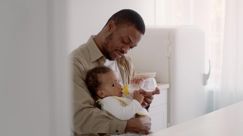 Caring african american father acting mom feeding his adorable curly baby son with milk bottle, carrying kid on hands at kitchen, tracking shot, slow motion Video Stok