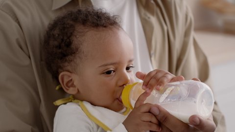 Baby feeding. Cute little curly african american baby drinking milk from kid bottle, feeling sleepy, caring daddy helping kid, close up portrait, slow motion