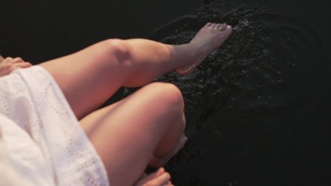 A woman wets her feet in the river at sunset sitting on the bridge. Close-up, slow motion.