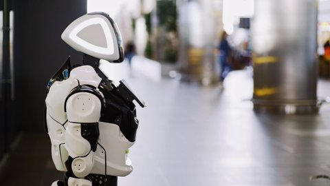 Black and white digital smart robot stands near column on airport waiting room territory against blurred tourists and sunlight reflections close view. Modern assistant