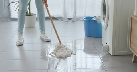 Broken washing machine leaking water on the floor and disappointed woman cleaning with a mop