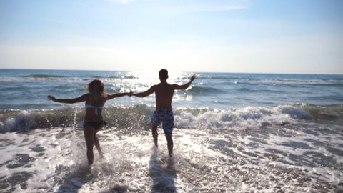 Young happy couple running at the beach to the sea and holding hands of each other. Girl and boy having fun together at summer vacation or holiday. Close up Rear back view Slow motion