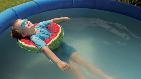 Pleasant baby boy smiling relaxing in watermelon safety buoy wearing goggles floating at inflatable rubber swimming pool happy male kid sunbathing enjoying outdoor summer vacation at sunny day