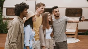 Group of young multicultural friends smiling and taking selfie on smartphone at camper site, happy multiethnic men and women having fun together while posing near motorhome in camping, sllow motion