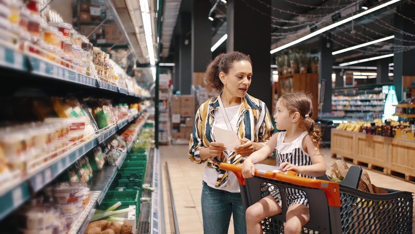 Little girl and her mom, housewife, enjoy family shopping together. A woman takes packed products from the shelf, wrapped mushrooms and puts them in a trolley. Royalty-Free Stock Footage #1075779143