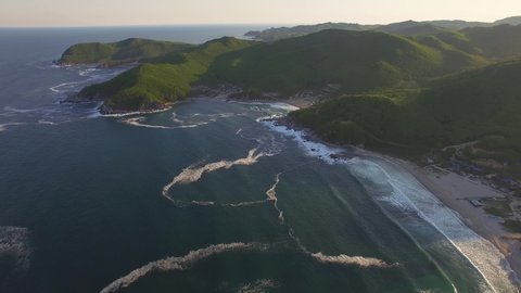 Drone view of the beautiful sea coast with clear blue water and rocky hills at sunset. The coast is covered with green vegetation. Foamy surf along the shore.