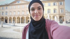 Tourist muslim woman makes a selfie video call holding mobile,phone point of view,arab girl wearing hijab smiling and looking at the camera with city in the background smartphone pov
