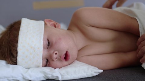 close-up view of a little child kid lying on the sofa with high temperature and cold compress on the forehead and mother's hands covering him with white blanket. cold flu illness care concept