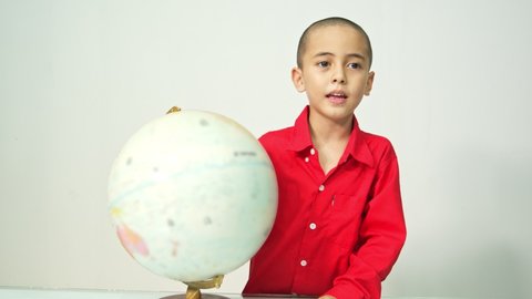 A boy with a skinhead in a red shirt is spinning the globe..A boy with skinhead hair in a red shirt is spinning a globe and .explaining about the world map in a white room..