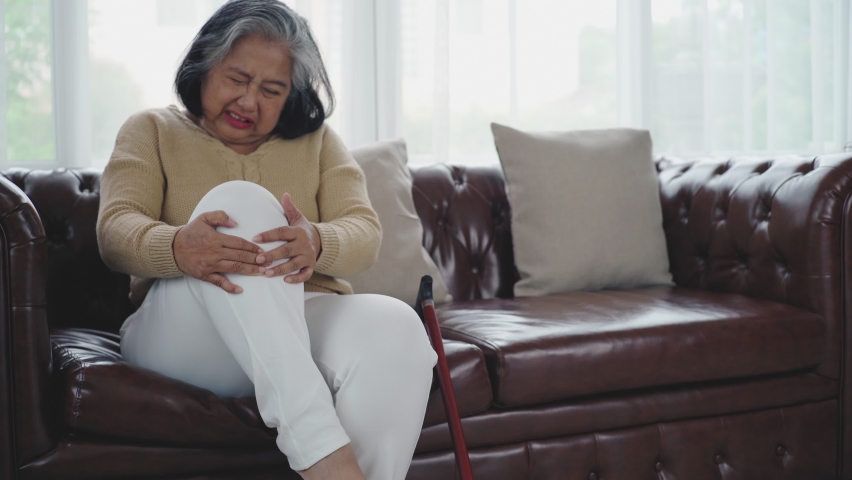 Senior adult with a painful expression from a serious legs and knee pain having difficulty getting up, sitting down on the sofa and moving around the house Royalty-Free Stock Footage #1075796108