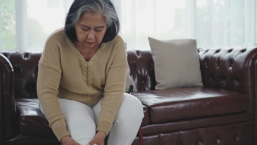 Senior adult with a painful expression from a serious legs and knee pain having difficulty getting up, sitting down on the sofa and moving around the house Royalty-Free Stock Footage #1075796120