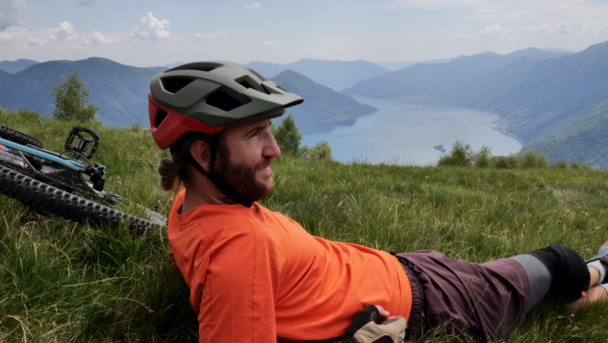 Slow motion: Mountain biker takes a moment on grassy knoll to appreciate mountain and lake scene