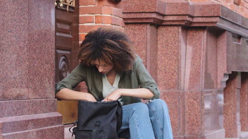 Young curly-haired African American girl sitting on steps outdoors nervously looking in her bag for keys, wallet or smartphone. Mixed Race black woman worried anxious scared because she lost things Royalty-Free Stock Footage #1075803929