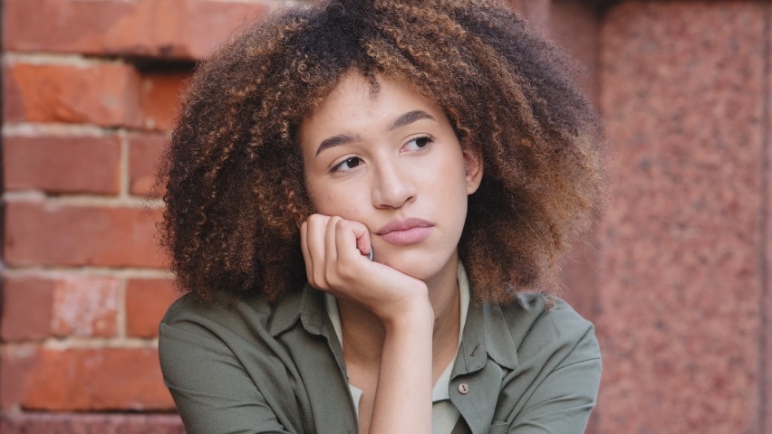 Closeup pensive young African American woman spend free time alone sitting outdoors lost deep in thoughts, feeling boredom. Looking depressed thinking about personal troubles, melancholic mood concept | Shutterstock HD Video #1075803932