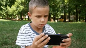 A boy of 6-7 years old with enthusiasm looks at the smartphone screen, plays a game on the phone. Park in the background, green trees