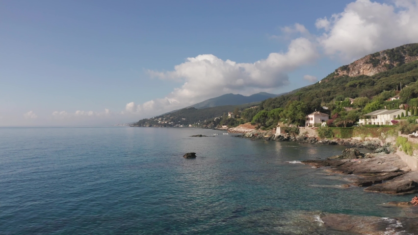 Aerial view of characteristic houses on coastline of corsica in a summer sunny day. Rocky shore,vivid blue sea, green vegetation on hill, trees, mountain in background