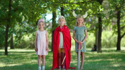 Three children in costumes looking at camera and waving hello in park. Boy wearing red cape and king crown and holding sword, girls in princess diadem and viking helmet. Kids role playing together