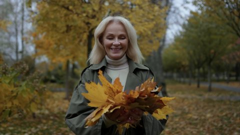 Old woman having fun at autumn park. Cheerful mature woman throwing yellow leaves with joyful mood outdoors. Portrait of happy blonde woman playing with foliage in autumn weather. 