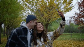 Funny young couple in love taking selfie on their mobile phone with autumn background. Close up of happy smiling lovers taking video on smartphone with cheerful faces in park. 