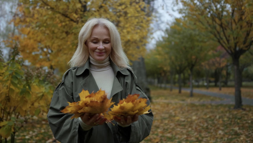Closeup of happy elderly lady having fun with foliage in autumn park. Cheerful senior woman throwing yellow leaves with enjoyment outdoors. Charming mature model relaxing in fall place.
