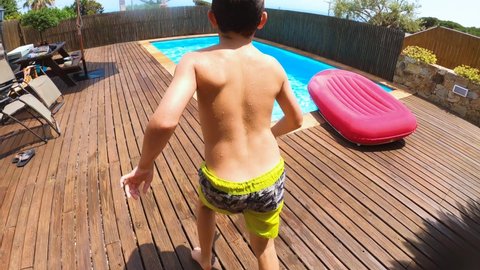 SLOW MOTION, UNDERWATER: Young boy speeding and jumping into swimming pool, kid doing a cannonball into clear water of a summer house. Courtyard of a summer house with wooden floor.