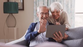 Happy senior couple using digital tablet for video conference and waving hand while laughing at home. Smiling grandparents making video call and greeting the family sitting on sofa.  - Powered by Shutterstock - Get 15% off with code: PIKWIZARD15
