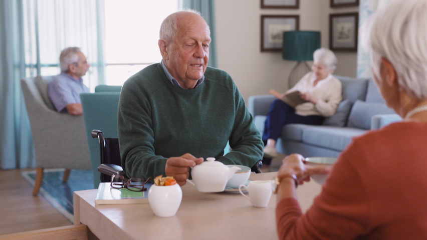 Smiling senior man pouring tea in cup from teapot in care facility with wife sitting at table in the common area. Elderly man in care centre sitting on wheelchair and serving tea. Royalty-Free Stock Footage #1075814267