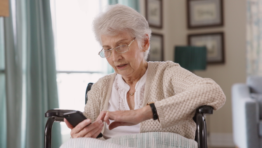 Elderly woman wearing spectacles sitting on wheelchair using smart phone. Happy old disabled woman messaging on cellphone at nursing home. Senior patient using mobile phone and smiling at camera.